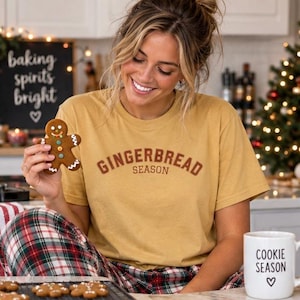 May include: A mustard yellow short-sleeve t-shirt featuring the text GINGERBREAD SEASON in brown arched lettering across the chest. The shirt is worn with red, green, and white plaid pants. A gingerbread man cookie is held in the foreground.