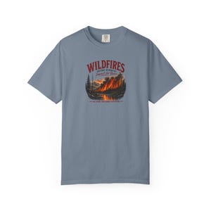 May include: A slate blue short-sleeve t-shirt featuring a graphic of a forest fire with the text "WILDFIRES DESTROY IN MINUTES IMPACT FOR YEARS" and "ONE SPARK CAN CHANGE EVERYTHING" printed in red and dark blue ink on the center chest area.