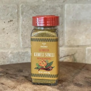Może przedstawiać: Szklany słoik przyprawy Khmeli Suneli z czerwoną zakrętką. Etykieta zawiera napis "Khomli Georgian Khmeli Suneli Seasoning" z ilustracją kwiatową i papryczką chili. Waga netto 50g.