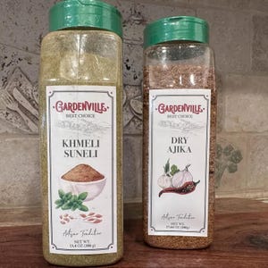 Może przedstawiać: Dwa przezroczyste plastikowe pojemniki na przyprawy z zielonymi pokrywkami. Jeden zawiera KHMELI SUNELI, drugi DRY AJIKA. Etykiety z logo "Gardenville Best Choice" i ilustracjami składników. KHMELI SUNELI ma 380 g, a DRY AJIKA ma 500g.