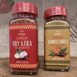 Może przedstawiać: Dwa szklane słoiki z gruzińską przyprawą. Jeden słoik ma czerwoną etykietę z napisem "DRY AJIKA SEASONING" i grafiką czosnku i papryczek chili. Drugi słoik ma beżową etykietę z napisem "KHMELI SUNELI SEASONING" i grafiką kwiatu i papryczki chili. Każdy słoik ma czerwoną pokrywkę.