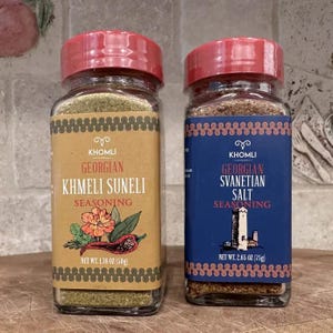 Może przedstawiać: Dwa szklane słoiki gruzińskich przypraw z czerwonymi pokrywkami. Jeden słoik jest oznaczony jako "Khmeli Suneli", a drugi jako "Svanetian Salt". Etykiety zawierają tekst i ilustracje kwiatów i wieży. Każdy słoik zawiera 50g i 75g przyprawy.