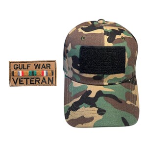 May include: Camouflage baseball cap with a black patch and a Gulf War Veteran patch. The cap features a woodland camouflage pattern in shades of green, brown, and black. The Gulf War Veteran patch is rectangular with text and colored stripes.
