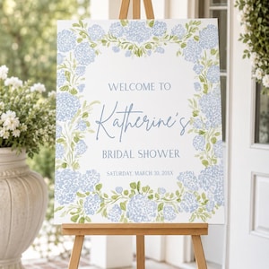 May include: A wooden easel displays a white sign with the text "Welcome to Katherine's Bridal Shower." The sign is decorated with a floral border of blue hydrangeas and green leaves. The date is Saturday, March 30, 20XX.