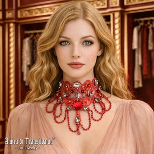 May include: A red lace choker necklace featuring intricate embroidery, dangling red beads, and clear faceted crystals. The design includes a central heart-shaped red stone and smaller circular red accents, creating a layered, ornate appearance.
