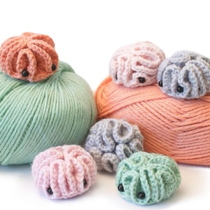 May include: A collection of colorful, crocheted brain toys in shades of peach, pink, gray, and green, each with black bead eyes. The toys are displayed on top of yarn skeins. The text "amigurumi brain toy crochet pattern" is at the top.