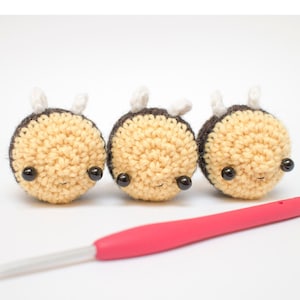 May include: Three crocheted bumblebees with black eyes and white wings. The bees are yellow and brown and are sitting on a white surface. A pink crochet hook is in the foreground.