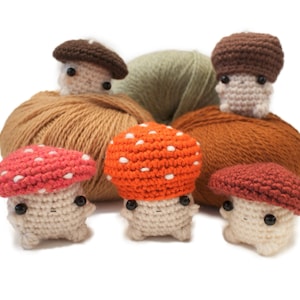 Puede incluir: Imagen de un patrón de ganchillo de setas amigurumi. La imagen presenta varias setas pequeñas tejidas a ganchillo con diferentes colores de sombrero, incluyendo rojo con puntos blancos, naranja con puntos blancos y marrón. El texto dice "mushroom no. 1 crochet pattern."
