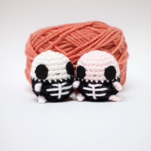 Op de afbeelding: Twee gehaakte skeletfiguren, een witte en een roze, met zwarte ogen en een zwart skeletontwerp. De figuren zitten voor een bol oranje garen.