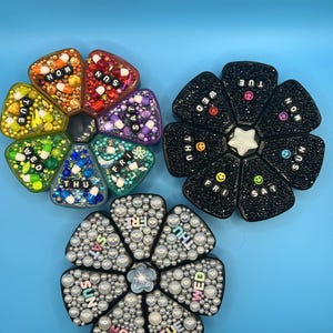 May include: Three decorative pill organizers in a flower shape. One is rainbow-colored with pills, one is black with white text and smiley faces, and the last is white with pearls and pastel text.