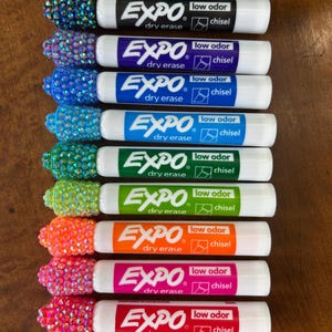 May include: An assortment of Expo dry erase markers with chisel tips. Each marker has a white barrel and a colored band indicating the ink color. The ends of the markers are embellished with colorful rhinestones. The markers are low odor.