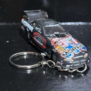 以下が含まれることがあります： 黒と赤のMopar Dodgeレーシングカーキーホルダー。車にはカラフルなグラフィックとMoparのロゴがあります。キーホルダーには、シルバーの金属リングとチェーンが付いています。