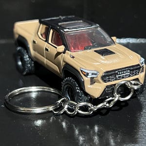 Puede incluir: Un llavero de camioneta Toyota Tacoma de color beige con techo negro, guardabarros negros y neumáticos todoterreno negros. La parrilla delantera muestra el logotipo de Toyota y una anilla de metal plateado está unida al parachoques delantero.