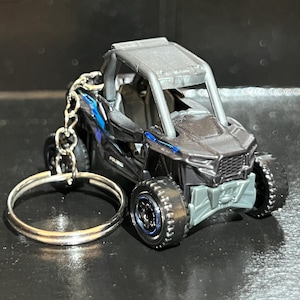 Könnte beinhalten: Ein schwarzer und grauer Spielzeug-Offroad-Fahrzeug-Schlüsselanhänger. Das Miniaturfahrzeug hat einen Überrollkäfig, große Reifen und einen silbernen Schlüsselring. Das Wort "POLARIS" ist auf der Seite des Fahrzeugs zu sehen.