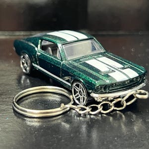Może przedstawiać: Brelok do kluczy w kształcie metalicznego, ciemnozielonego samochodzika z dwoma białymi pasami biegnącymi przez maskę i dach. Miniaturowy pojazd ma srebrne koła, przezroczystą przednią szybę oraz metalowe kółko przymocowane krótkim łańcuszkiem.