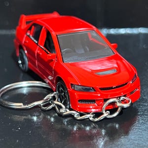 Puede incluir: Un llavero de coche modelo rojo. El coche en miniatura tiene un alerón, ruedas negras y un interior detallado. El llavero está sujeto con una cadena y un anillo plateados.