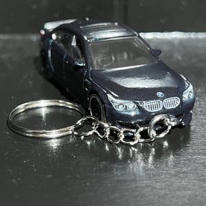 May include: A dark blue BMW sedan model car keychain featuring a metallic finish, a silver split ring, and a short chain. The miniature vehicle displays a detailed front grille, headlights, and the BMW logo on the hood, with a sunroof visible.