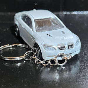 May include: A white BMW model car keychain featuring a detailed front grille, headlights, and hood vents. The miniature vehicle is attached to a circular silver metal split ring by a short silver metal chain, showcasing a realistic automotive design.