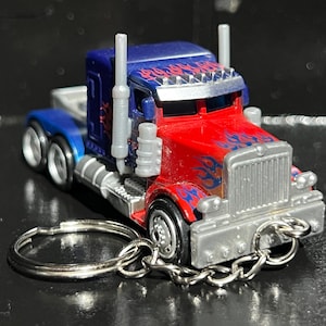 May include: A miniature red, blue, and silver semi-truck toy with flame decals, attached to a keychain. The truck has a detailed front grill and exhaust pipes. The wheels are black and silver.