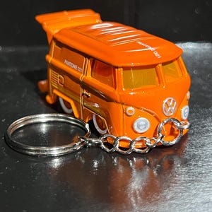 May include: Orange Volkswagen van keyring with the text "PANTONE 021". The van has white tyres and a silver keyring ring. The van has a wing on the top.