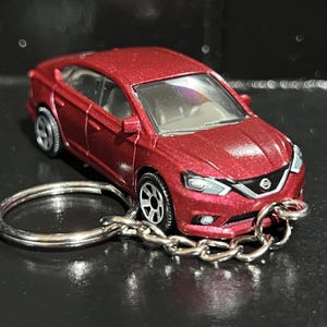 May include: A miniature red car keyring. The car has a detailed design with silver wheels and a black interior. The keyring includes a silver key ring and chain. The car is approximately 7.6 cm long.