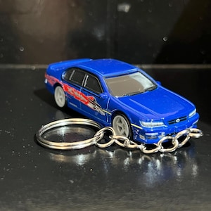 May include: A blue die-cast model car keychain. The car has white wheels and red and orange graphics on the side. The keychain has a silver chain and a silver key ring.
