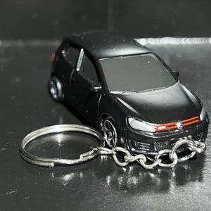 May include: A black die-cast model car keyring. The miniature car has a red stripe on the front grill and silver wheels. The keyring ring and chain are silver. The car is a hatchback style.