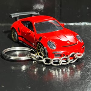 May include: A red toy sports car keyring with gold-coloured rims and a silver chain. The car has a spoiler and is on a silver key ring. The car is a miniature replica of a Porsche.