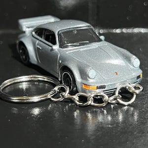 May include: Silver die-cast model car keyring. The miniature car has a spoiler and orange-tinted headlights. The keyring has a silver ring and chain. The car is approximately 5 cm long.