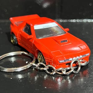 Puede incluir: Un llavero de coche deportivo rojo brillante de metal fundido a presión con alerón trasero, toma de aire en el capó y ruedas doradas. El parabrisas tiene una banda blanca con texto y el parachoques delantero incluye cuatro luces circulares.
