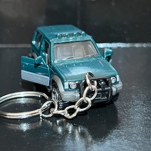 Puede incluir: Un llavero de SUV en miniatura de color verde azulado. El coche de juguete tiene un diseño detallado, que incluye faros, un parachoques delantero y una puerta abierta. El llavero es plateado y está sujeto a la parte delantera del vehículo.