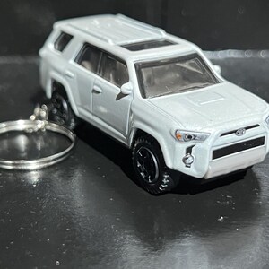 May include: A white Toyota 4Runner toy SUV keychain featuring black wheels, a detailed front grille with the Toyota emblem, clear headlights, and a silver metal split ring attached to the rear side of the vehicle for secure key storage.