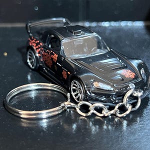 May include: Black die-cast model car keychain with red floral accents. The miniature car features a spoiler and detailed wheels. The keychain includes a silver metal ring and chain. A collectible item.