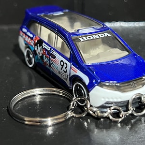 Op de afbeelding: Een blauw-wit Honda minivan speelgoed met het woord "HONDA" op de motorkap. Het speelgoed heeft het nummer "93" en de woorden "BOOST TIME!" aan de zijkant. Een zilveren sleutelhanger is aan het speelgoed bevestigd.