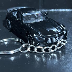 May include: A black die-cast model car keychain. The miniature car features a detailed design with a spoiler and a silver chain and keyring. The car has a prominent front grille and headlights.