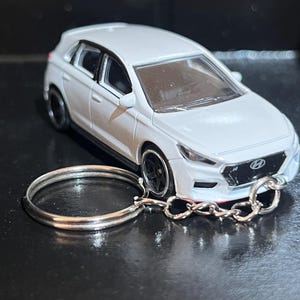 May include: A white Hyundai i30 toy car keyring. The miniature car has black wheels and a detailed design. The keyring includes a silver key ring and chain. The car is a hatchback model.
