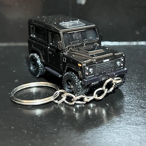 Puede incluir: Un llavero de SUV en miniatura negro con una cadena y un llavero plateados. El coche de juguete tiene detalles, incluyendo un techo solar y neumáticos todoterreno. El llavero es una pequeña y detallada réplica de un vehículo clásico.