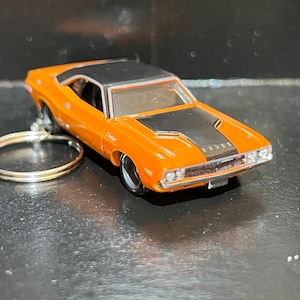 May include: A die-cast metal Dodge Challenger toy car keychain featuring a bright orange body with a black roof and a black hood stripe. The vehicle has clear windows, silver-toned headlights, a front grille, and a circular metal split ring attached.