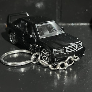 May include: A black die-cast model car keychain. The miniature car features detailed design elements, including wheels, windows, and a rear spoiler. The keychain has a silver metal ring and chain.