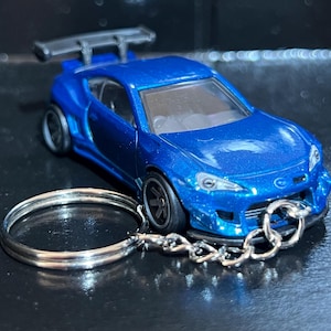 May include: A blue die-cast model car with a rear spoiler and black wheels. The car is attached to a silver keychain. The car's body has a metallic finish.