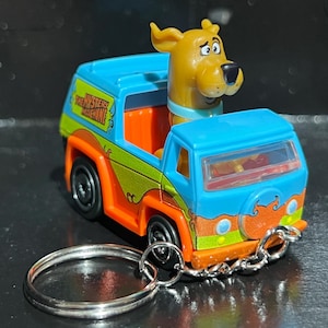 May include: A miniature toy Mystery Machine van from Scooby-Doo, with a blue roof, orange and green sides, and a Scooby-Doo figure. Includes a silver keychain. The van has the words "The Mystery Machine" on the side.