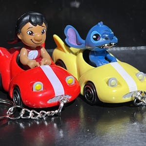 以下が含まれることがあります： 小さなミニカーに乗ったリロとスティッチのキャラクターキーホルダー2個セット。リロのフィギュアは白いレーシングストライプが入った赤い車に、スティッチのフィギュアは白いレーシングストライプが入った黄色い車に乗っている。どちらの車もフロントバンパーに銀色の金属製キーリングが付いている。