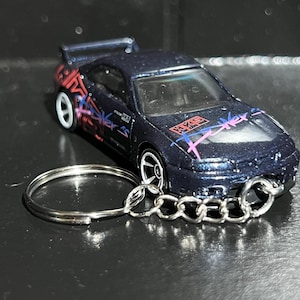 May include: A dark blue metallic toy car keychain featuring a rear spoiler, white wheels, and pink and blue graphic decals on the hood and sides. The car includes the text HKS printed on the hood and is attached to a silver metal split ring.