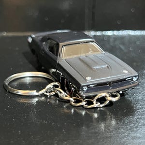 May include: A matte black die-cast muscle car keychain featuring a detailed hood, front grille, and headlights. The miniature vehicle is attached to a standard silver-toned metal split ring by a short, sturdy silver-colored metal chain link.