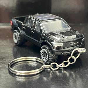 Puede incluir: Un vehículo de juguete tipo camioneta Ram de color negro unido a un llavero de metal plateado. La camioneta cuenta con un diseño de cabina doble, una parrilla frontal detallada con el logo RAM, ventanas tintadas, techo solar y ruedas todoterreno.