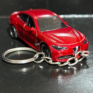 May include: A red die-cast model car keyring featuring a detailed saloon design with black wheels, a triangular front grille, and a silver metal split ring attached to the front bumper by a short silver chain, showcasing a glossy paint finish.