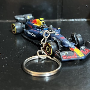 Puede incluir: Un llavero de coche de carreras de Fórmula 1 en miniatura. El coche es principalmente azul marino con detalles rojos y amarillos, con el logotipo "Oracle". El llavero incluye un llavero y una cadena plateados.