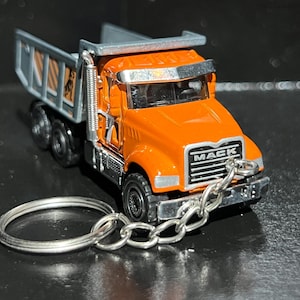 Op de afbeelding: Oranje Mack dumper speelgoed met een zilveren sleutelhanger. De vrachtwagen heeft zwarte wielen, een zilveren grille met het woord "MACK" en een zilveren laadbak. De sleutelhanger is gemaakt van zilver metaal.