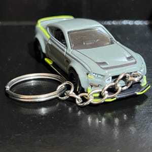 Puede incluir: Un llavero de coche de juguete gris con ruedas negras y detalles en verde neón. El coche tiene una cadena y un llavero plateados. El coche tiene un alerón y franjas de carreras.