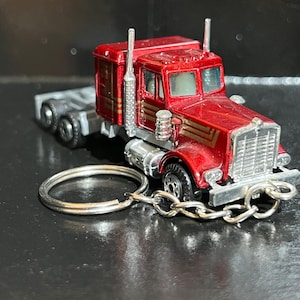 Op de afbeelding: Een rode en gouden speelgoed vrachtwagen sleutelhanger. De miniatuur vrachtwagen heeft een gedetailleerd ontwerp met een zilveren grille, uitlaatpijpen en een trekhaak. De sleutelhanger bevat een zilveren ketting en ring.